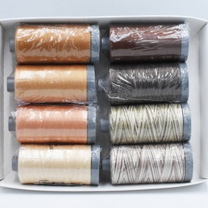 May include: A collection of eight thread spools in a white box. The colours vary from light beige to dark brown. Each spool is encased in clear plastic. The spools are arranged in two rows of four.