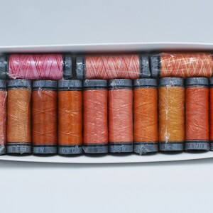 May include: A collection of thread spools in a white box. The spools are in various shades of pink, orange, and yellow, and are wrapped in clear plastic. The thread is likely cotton, suitable for sewing and craft projects.
