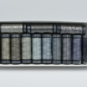 May include: A selection of thread spools in a range of grey and blue hues, presented in a white box. The spools are neatly arranged, with the thread wound around them. Each spool is labelled with text.