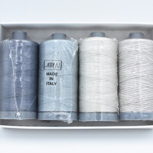 May include: Four spools of thread in a white box, in shades of grey and white. The spools are wrapped in clear plastic. One spool has the text "MADE IN ITALY". The thread is suitable for sewing projects.