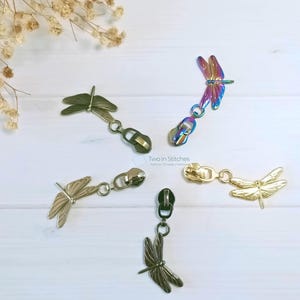 May include: Five metal dragonfly zipper pulls in gold, iridescent, and bronze. Each pull is attached to a dragonfly charm with a small metal ring. The text "Two in Stitches" is visible. These are ideal for craft projects.