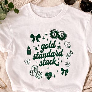 May include: White t-shirt with a green graphic design. The design includes the text "gold standard stack" with various symbols like dice, shamrocks, and bows. Two 8-ball pool balls are also featured in the design.