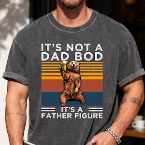May include: Dark gray t-shirt with the text "IT'S NOT A DAD BOD IT'S A FATHER FIGURE" and a graphic of a bear holding a beer. The bear is brown and stands in front of a retro sunset design.