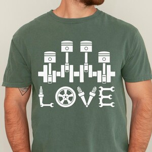May include: Moss green t-shirt with a white graphic of engine parts spelling "LOVE". The design includes pistons, a spark plug, a wheel, and wrenches. The shirt is a Comfort Colors brand.