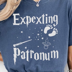 May include: A blue t-shirt with the text "Expecting Patronum" in white. The design includes a white wand, a baby pacifier, and two white footprints.