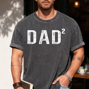 May include: Dark gray t-shirt with the white text "DAD²". The shirt has rolled-up sleeves and a slightly faded appearance. The t-shirt is a casual style.