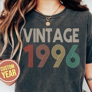 May include: Dark gray Comfort Colors t-shirt with the word "VINTAGE" in cream-colored, retro font. Below, the year "1996" is printed in red, yellow, and green. The shirt has a custom year option and is labeled "Pepper".