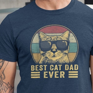 May include: A navy blue t-shirt with a retro-style graphic of a cat wearing sunglasses. The graphic is surrounded by a red, white, and yellow circle. The text "Best Cat Dad Ever" is printed below the graphic.