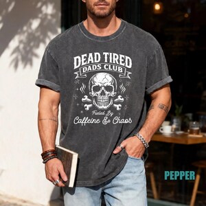 May include: Dark gray t-shirt with rolled sleeves featuring a skull and crossbones graphic. The text reads "DEAD TIRED DADS CLUB" above and "Fueled By Caffeine & Chaos" below. The shirt is worn by a person.