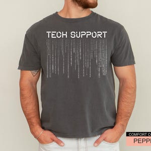 K&ouml;nnte beinhalten: Ein dunkelgraues T-Shirt mit den Worten "TECH SUPPORT" in wei&szlig;en Blockbuchstaben &uuml;ber einem vertikalen Bin&auml;rcode-Design. Das Shirt ist von der Marke Comfort Colors in der Farbe Pepper.