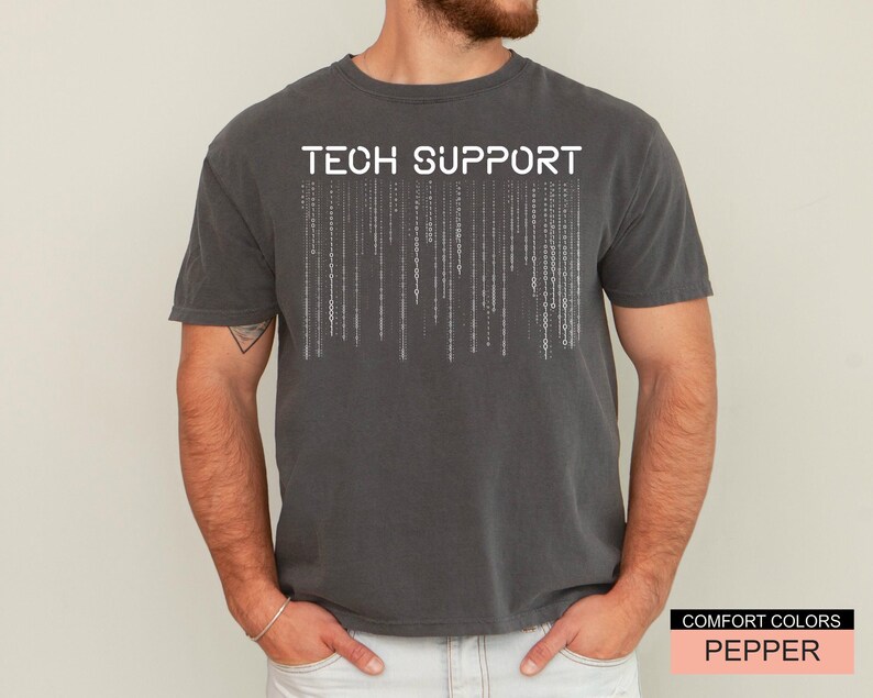 K&ouml;nnte beinhalten: Ein dunkelgraues T-Shirt mit den Worten "TECH SUPPORT" in wei&szlig;en Blockbuchstaben &uuml;ber einem vertikalen Bin&auml;rcode-Design. Das Shirt ist von der Marke Comfort Colors in der Farbe Pepper.