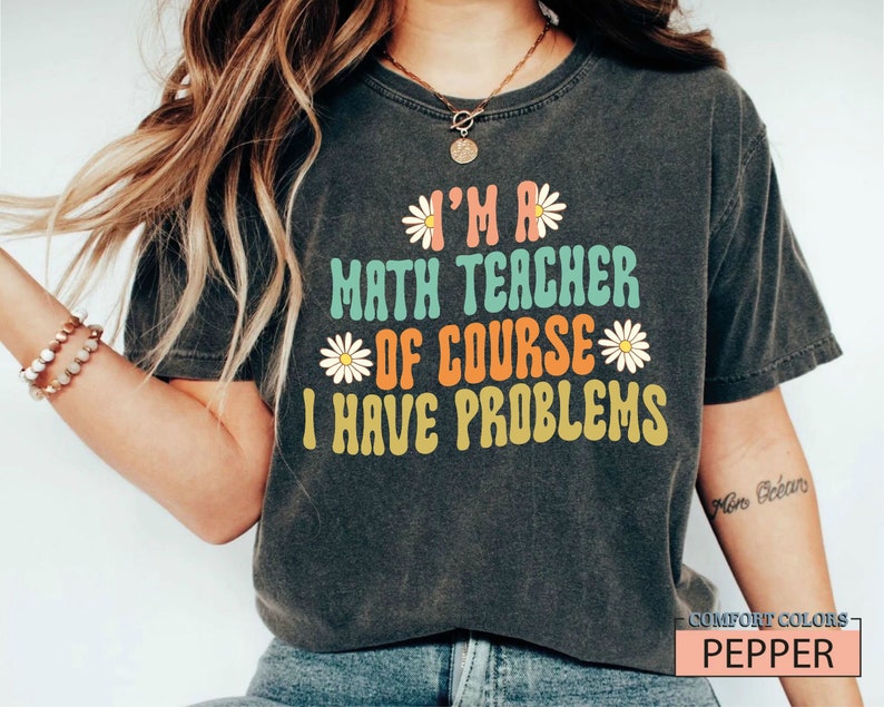 Pode incluir: Uma camiseta cinza escura com um gr&aacute;fico de estilo retr&ocirc; que diz "Eu sou professor de matem&aacute;tica, claro que tenho problemas". O gr&aacute;fico apresenta margaritas coloridas e uma fonte vintage.