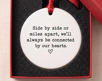 May include: A round white ceramic ornament featuring the black text: Side by side or miles apart, we'll always be connected by our hearts, with a small heart icon below. The ornament includes a red ribbon and rests in a red gift box.