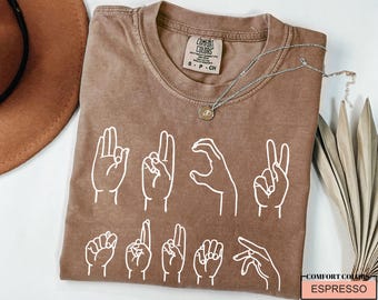 May include: A brown t-shirt with a white graphic of hands signing the word "love". The shirt has a distressed look and is made of a soft, comfortable fabric. The text "COMFORT COLORS ESPRESSO" is printed on the bottom right corner of the shirt.