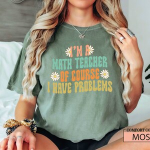Pode incluir: Uma camiseta verde com o texto "I'm a math teacher of course I have problems" em uma fonte retr&ocirc; com gr&aacute;ficos de margaridas. A camiseta est&aacute; em uma pessoa com cabelo longo e loiro.