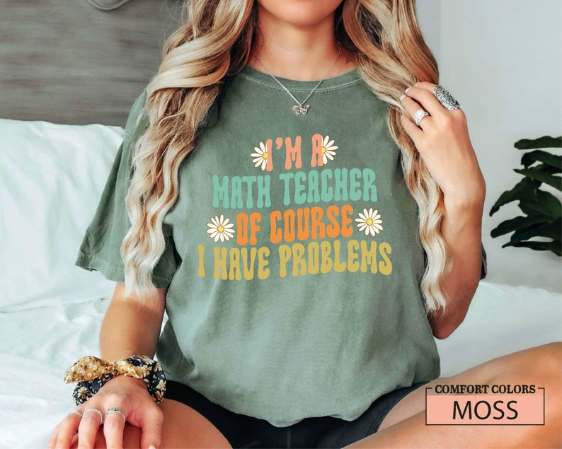Pode incluir: Uma camiseta verde com o texto "I'm a math teacher of course I have problems" em uma fonte retr&ocirc; com gr&aacute;ficos de margaridas. A camiseta est&aacute; em uma pessoa com cabelo longo e loiro.