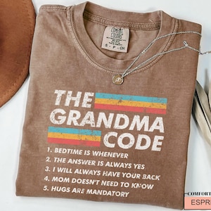 May include: A brown t-shirt with a vintage-style graphic that says "The Grandma Code" with a rainbow stripe design. The text below the graphic lists five rules of the Grandma Code: "Bedtime is whenever", "The answer is always yes", "I will always have your back", "Mom doesn't need to know", and "Hugs are mandatory".