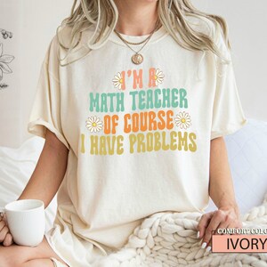 Pode incluir: Uma camiseta branca com o texto "I'm a math teacher of course I have problems" em uma fonte retr&ocirc; com detalhes de margaridas. A camiseta est&aacute; sendo usada por uma modelo e o texto "COMFORT COLORS IVORY" est&aacute; no canto inferior direito.