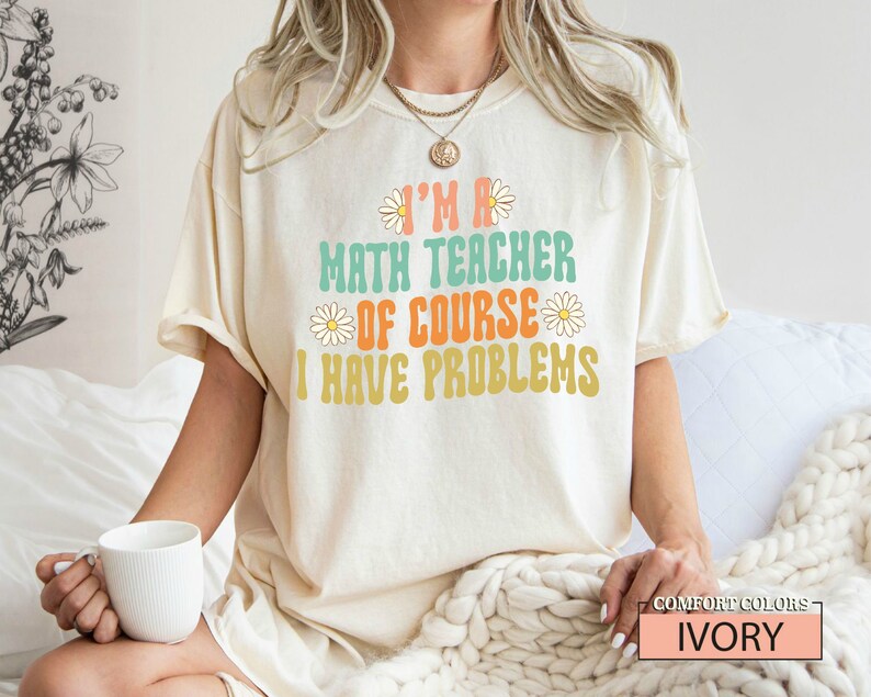 Pode incluir: Uma camiseta branca com o texto "I'm a math teacher of course I have problems" em uma fonte retr&ocirc; com detalhes de margaridas. A camiseta est&aacute; sendo usada por uma modelo e o texto "COMFORT COLORS IVORY" est&aacute; no canto inferior direito.