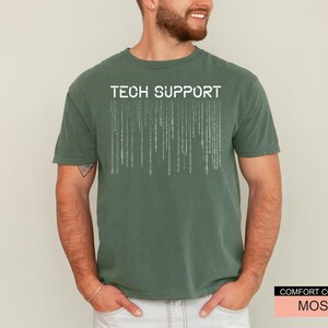 K&ouml;nnte beinhalten: Ein moosgr&uuml;nes T-Shirt mit dem wei&szlig;en Schriftzug "TECH SUPPORT". Unter dem Text befindet sich ein vertikales Matrix-Design. Das Shirt ist von der Marke Comfort Colors.