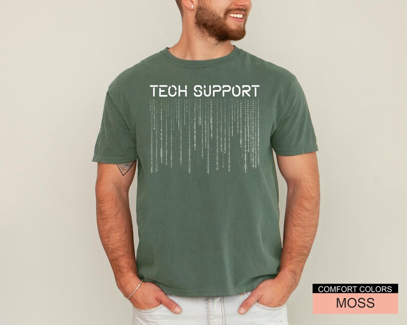 K&ouml;nnte beinhalten: Ein moosgr&uuml;nes T-Shirt mit dem wei&szlig;en Schriftzug "TECH SUPPORT". Unter dem Text befindet sich ein vertikales Matrix-Design. Das Shirt ist von der Marke Comfort Colors.