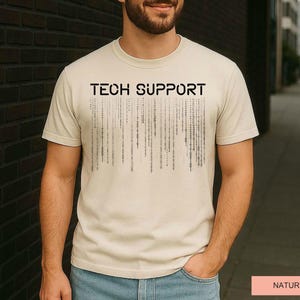 K&ouml;nnte beinhalten: Beiges T-Shirt mit dem schwarzen Aufdruck "TECH SUPPORT" auf der Brust. Darunter befindet sich eine Grafik aus vertikalen Linien, die an Bin&auml;rcode erinnern. Das Wort "NATUREL" ist auf einem rosa Rechteck gedruckt.