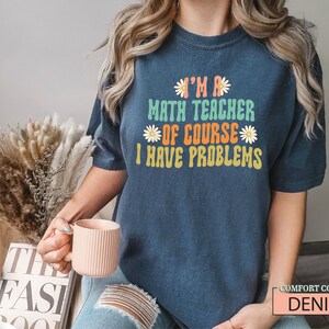 Pode incluir: Uma camiseta azul marinho com o texto "I'm a math teacher of course I have problems" em uma fonte retr&ocirc; colorida com detalhes de margaridas. A camiseta est&aacute; sendo usada por uma pessoa com cabelo longo e loiro.