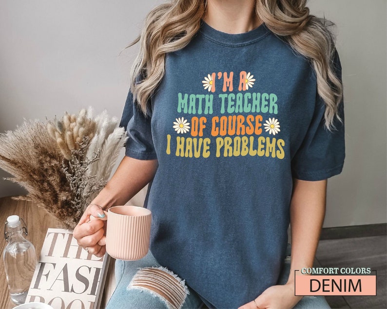 Pode incluir: Uma camiseta azul marinho com o texto "I'm a math teacher of course I have problems" em uma fonte retr&ocirc; colorida com detalhes de margaridas. A camiseta est&aacute; sendo usada por uma pessoa com cabelo longo e loiro.