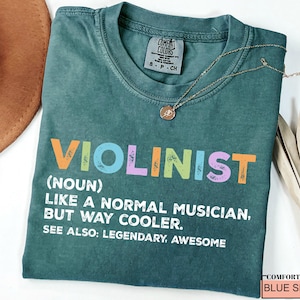 May include: A teal-colored t-shirt with the word "VIOLINIST" in colorful letters. Below the word is the definition: "(NOUN) LIKE A NORMAL MUSICIAN, BUT WAY COOLER. SEE ALSO: LEGENDARY, AWESOME." A gold necklace rests on the shirt.