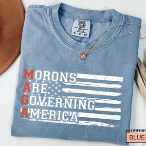 May include: A blue jean t-shirt with a white distressed American flag design and the text "MORONS ARE GOVERNING AMERICA" in white with the "A" in "AMERICA" in red.