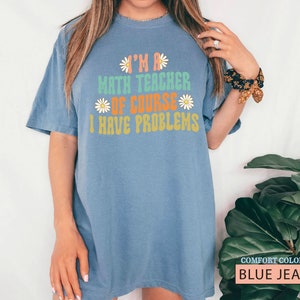 Pode incluir: Uma camiseta azul jeans com o texto "I'm a math teacher of course I have problems" em uma fonte retr&ocirc; com detalhes de margaridas.