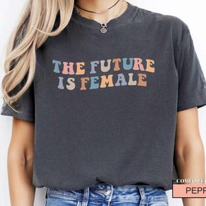 May include: A dark gray t-shirt with the text "The Future is Female" in rainbow colors. The text is in a retro font style.