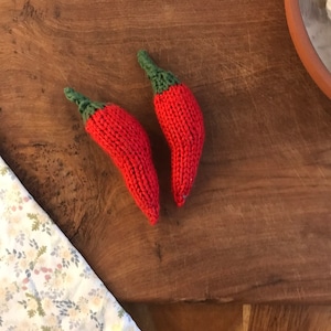 May include: Two red knitted chili peppers with green tops, lying on a wooden cutting board.
