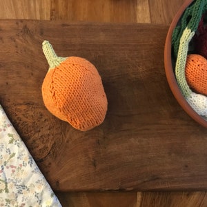 May include: An orange knitted pumpkin with a green stem sits on a wooden surface. A bowl filled with knitted vegetables, including carrots, onions, and beets, is visible. A floral patterned cloth is in the corner.