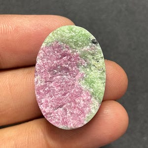 Peut inclure: Une pierre précieuse cabochon de forme ovale présentant une surface texturée avec une zone proéminente de matériau cristallin rose vif contrastant avec une matrice minérale vert clair, incluant une petite inclusion noire près du bord.