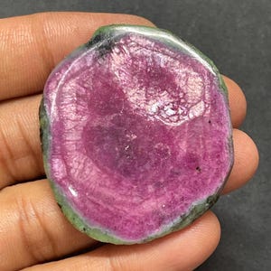 May include: A polished, circular gemstone slice featuring a vibrant magenta and deep pink center with a distinct, textured crystalline structure. The outer edge is bordered by a thin, irregular band of muted green and grey mineral material.