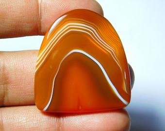 May include: A polished, orange agate gemstone with white banding. The stone has a rounded top and a flat bottom, with a smooth, glossy surface. The bands create a wave-like pattern across the surface of the stone. The gemstone is held in a person's hand.