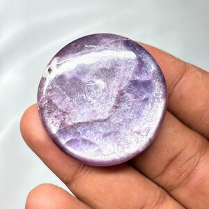 K&ouml;nnte beinhalten: Ein runder, polierter Edelstein mit einem wirbelnden Muster aus Lavendel und Wei&szlig;. Der Stein wird in einer Hand gehalten, wodurch seine glatte, reflektierende Oberfl&auml;che hervorgehoben wird. Die Farbe des Edelsteins reicht von hell- bis tiefviolett.