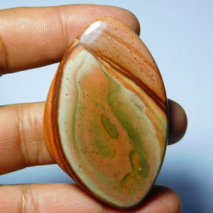 May include: A polished, teardrop-shaped gemstone with a swirling pattern of orange, brown, green, and cream colors. The stone is held in a hand, showcasing its smooth, glossy surface. This natural stone is ideal for jewelry making or collecting.