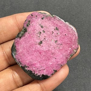 May include: A flat, irregularly shaped slice of pink ruby gemstone with a rough, dark, and textured outer edge. The surface of the stone displays a vibrant magenta hue with scattered black mineral inclusions throughout the crystalline structure.