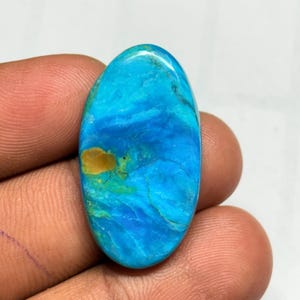 Op de afbeelding: Een ovaalvormige edelsteen met een levendige mix van blauwe en turkoois tinten, geaccentueerd door een goudbruine vlek. Het gepolijste oppervlak toont natuurlijke patronen, wat een unieke uitstraling creëert. Ideaal voor het maken van sieraden.