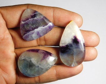 May include: Three polished fluorite gemstones in shades of purple, white, and blue. The gemstones are teardrop and triangular shaped. The stones are held in a hand against a white background. The gemstones have a translucent appearance.