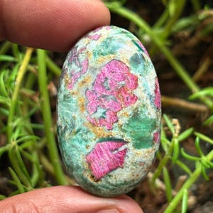Peut inclure: Pierre précieuse de forme ovale avec un mélange de couleurs. La base est verte et grise, avec des inclusions roses et dorées vives. La surface polie est lisse, reflétant la lumière et mettant en valeur les motifs et textures naturels.