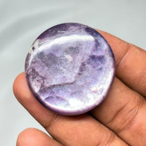 K&ouml;nnte beinhalten: Ein runder, polierter Edelstein mit einem wirbelnden Muster aus Lavendel und Wei&szlig;. Der Stein wird in einer Hand gehalten, wodurch seine glatte, reflektierende Oberfl&auml;che hervorgehoben wird. Die Farbe des Edelsteins reicht von hell- bis tiefviolett.