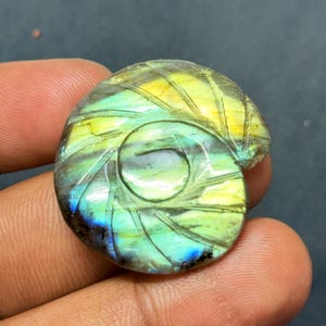May include: A carved labradorite gemstone in a spiral seashell design. The stone features iridescent colors of yellow, green, and blue. The gemstone is round and has a polished finish, showcasing the intricate carving details.
