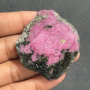 May include: A raw, irregular mineral specimen featuring a vibrant, textured magenta-pink center surrounded by a dark, metallic, and crystalline border. The stone displays a rough, natural surface with visible mineral inclusions and varied color contrast.