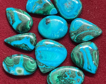 May include: A collection of polished gemstones with a mix of turquoise and green hues. The stones vary in shape, from rounded to teardrop, and display intricate patterns. The gemstones are arranged on a red surface.