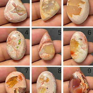Puede incluir: Una colección de nueve piedras preciosas cabujón pulidas, cada una con una matriz única de tonos crema, beige y naranja con inclusiones de ópalo. Las piedras tienen formas ovaladas y de lágrima, mostrando patrones minerales naturales.