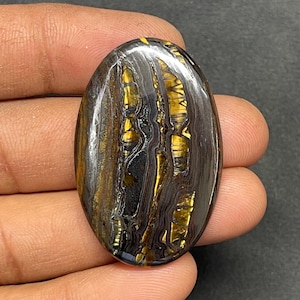 May include: Oval-shaped gemstone with a polished surface. The stone features a striking pattern of gold, black, and gray hues, with a layered, striped appearance. The gemstone is held in a hand, showcasing its size and detail.