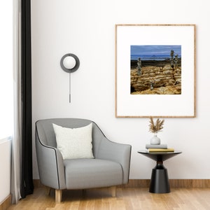 May include: A grey armchair with a white cushion sits in a living room with a black side table and a framed photograph of a rocky beach with stacked stones.